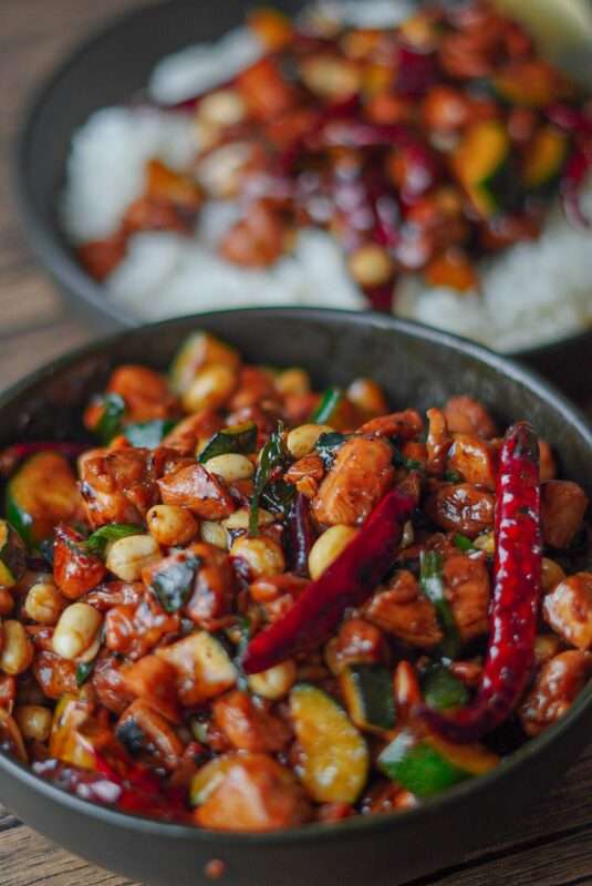 A colorful dish of kung pao chicken made out of red peppers, zucchini, green onions, stir fried chicken and brown sauce.