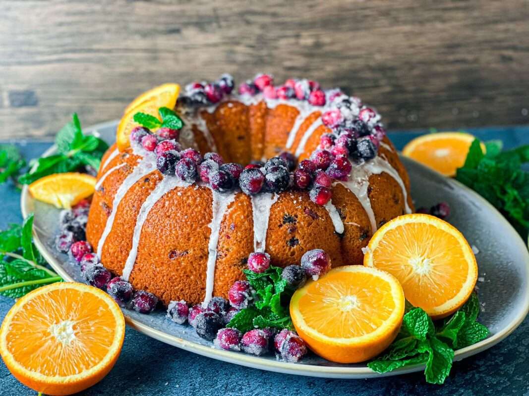 Perfect Cranberry Orange Bundt Cake on a white plate garnished with orange glaze and fresh fruits with 3 orange halves near it