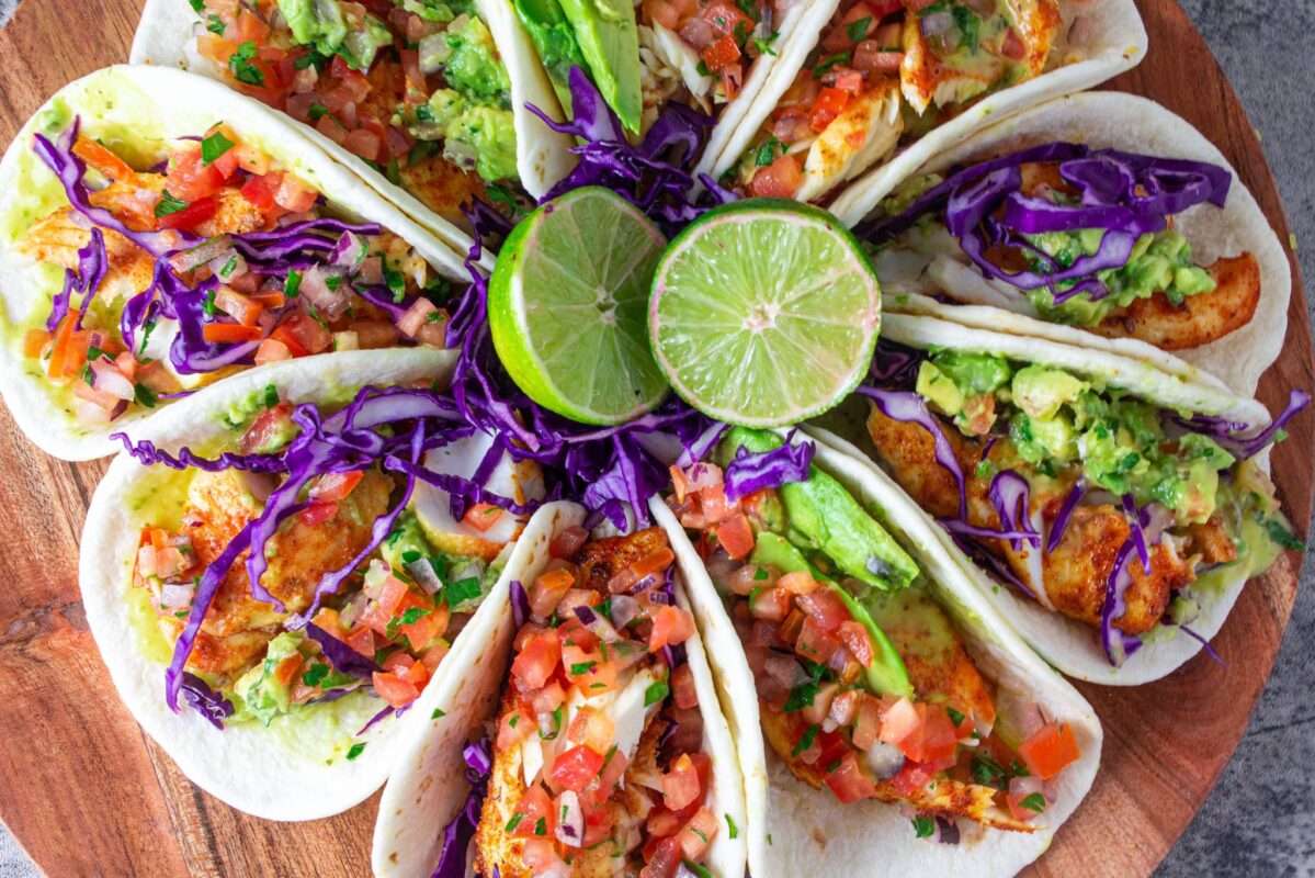 A flowery bunch of fish tacos served on a plate decorated with chopped vegetables and two halves of lemon.