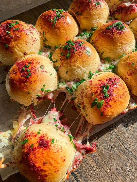 A group of golden slider buns are stuffed with gooey mozzarella cheese and musakhan slices. They are sprinkled with the lemony sumac powder.