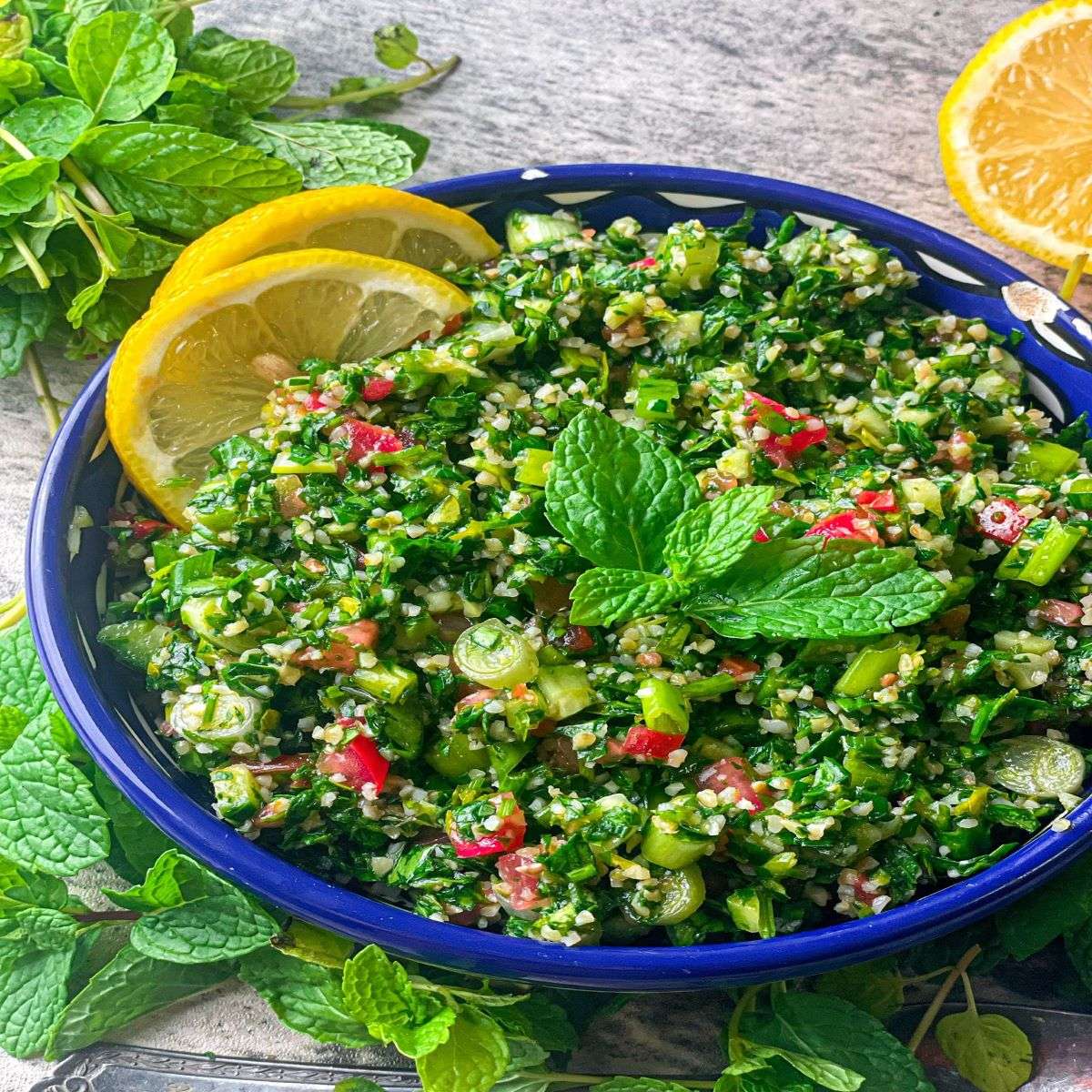 A delicious bowl of tabouli salad made of fresh parsley, burgul, and other ingredients. It is decorated with mint leaves and lemon slices.