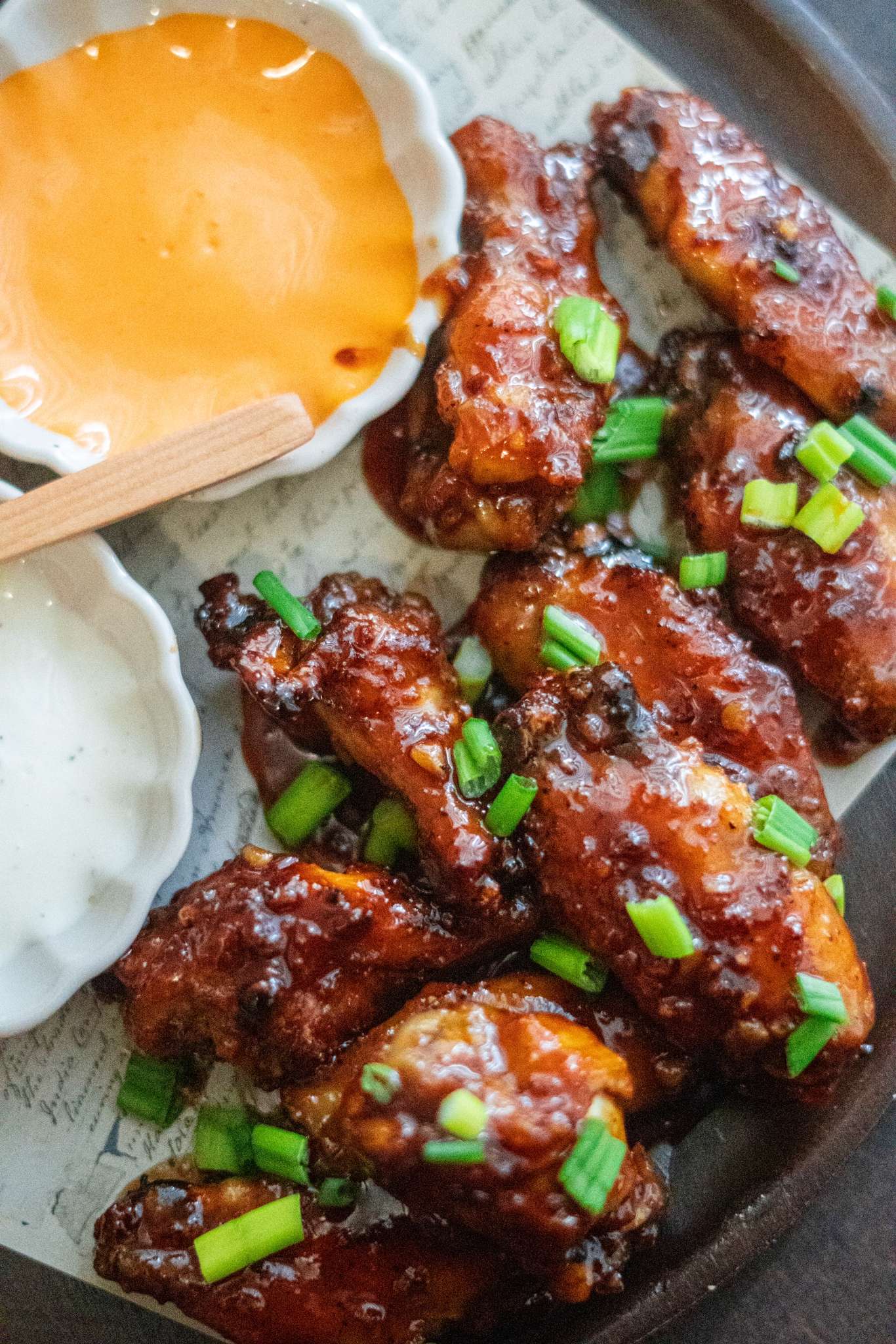 A plate of honey garlic siracha wings topped with fresh spring onions and served with a side of sauces.