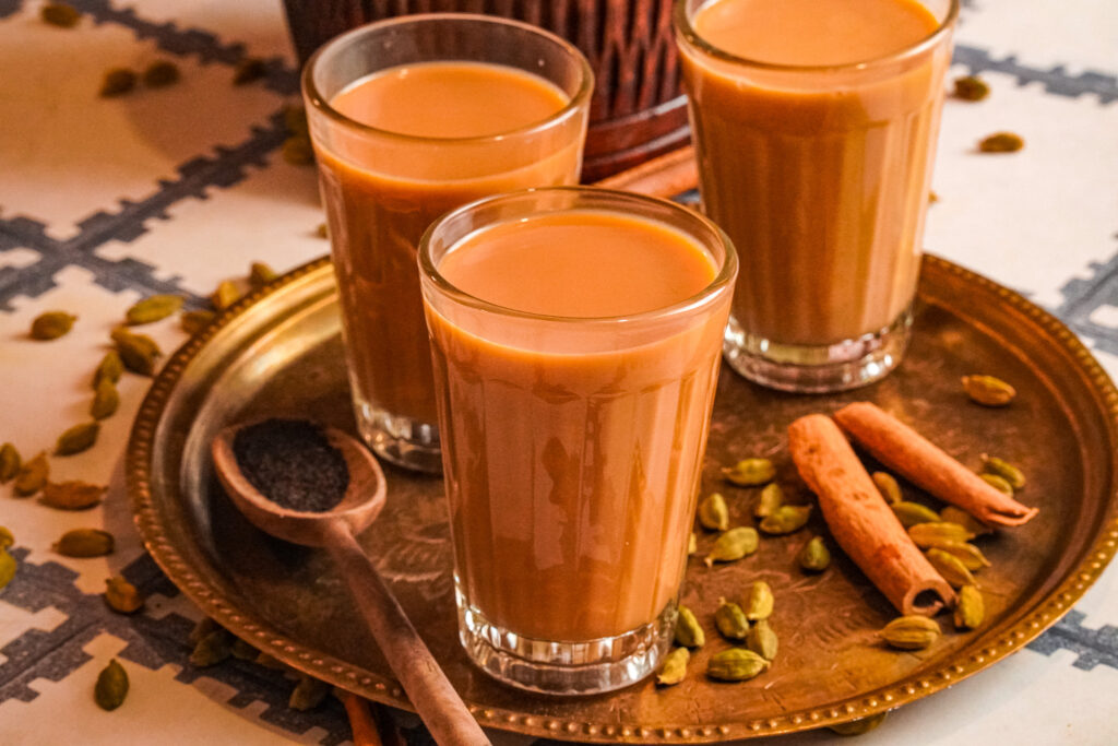 A tray serving three cups of hot karak chai with some cardamom and cinnamon