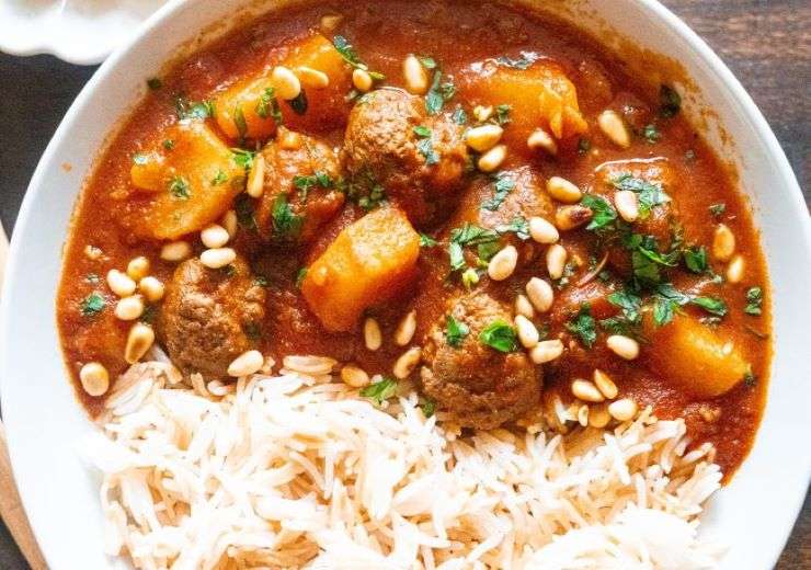 In a white plate, kufta balls are combined with chopped potatoes in a tomato sauce mixture. Near them, there is vermicelli rice in the same plate.