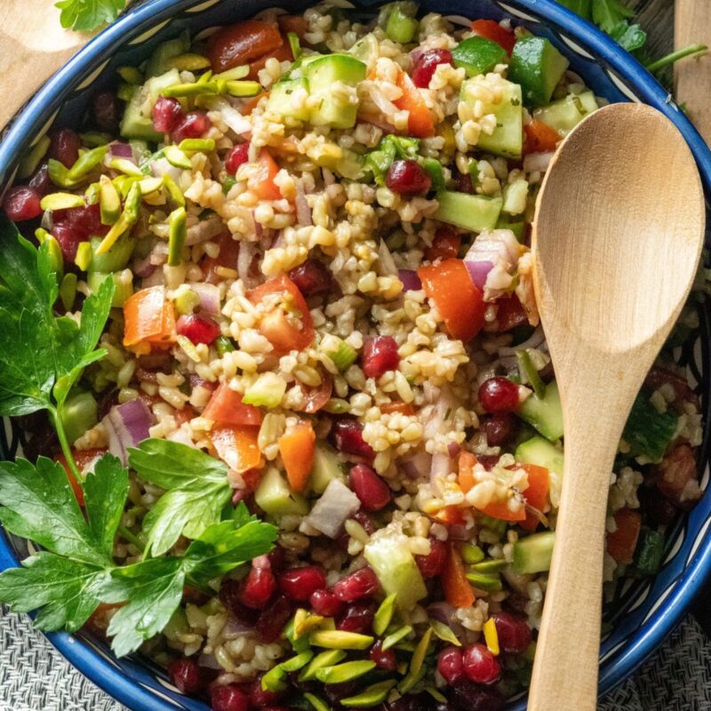 a colorful and nutritious freekeh salad presented on the table with fresh parsley, tomatoes, red onions, and cucumbers, topped with pomegranate seeds and thinly sliced pistachios.