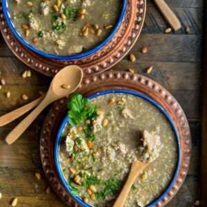 Two bowls of freekeh soup showing cilantro and pine nuts and a spoon scooping a chunk of chicken.