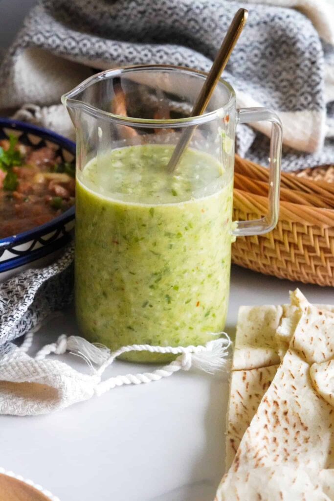 Tatbeeleh (Shatta) in a jar with a spoon inside of it, Pita bread next to it. Baskets behind the jar.