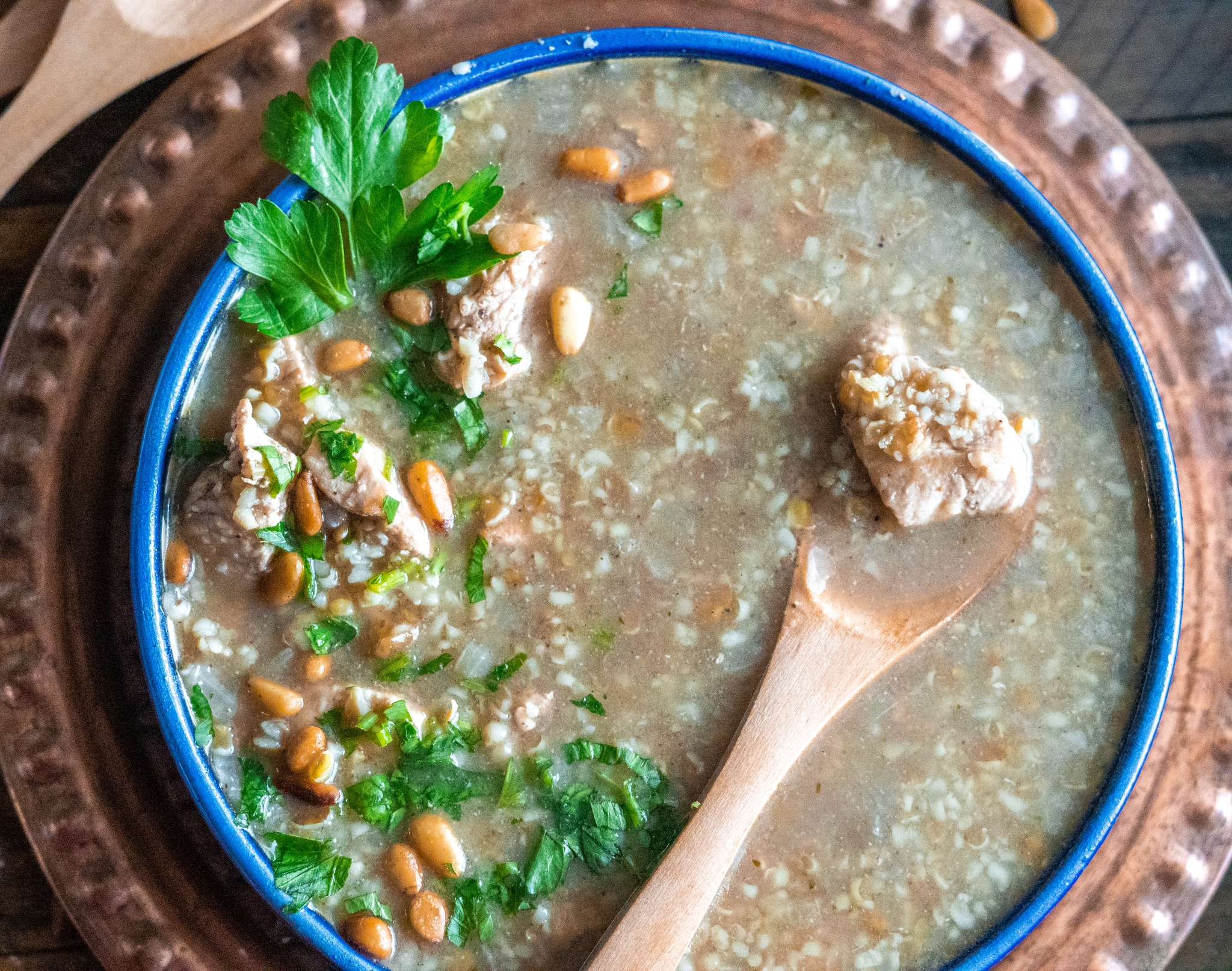 A bowl of freekeh soup showing cilantro and pine nuts and a spoon scooping a chunk of chicken.