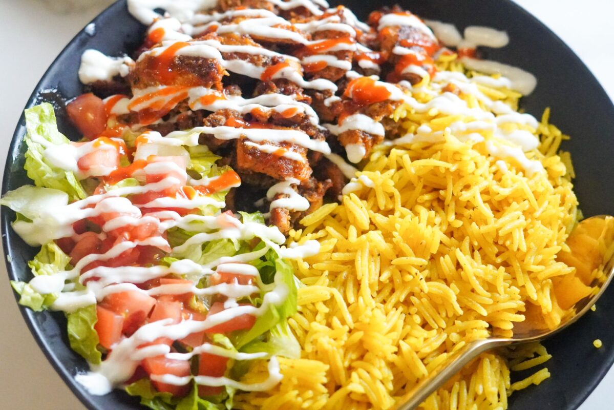 a dish of juicy chicken served with yellow rice, chopped vegetables, and white sauce