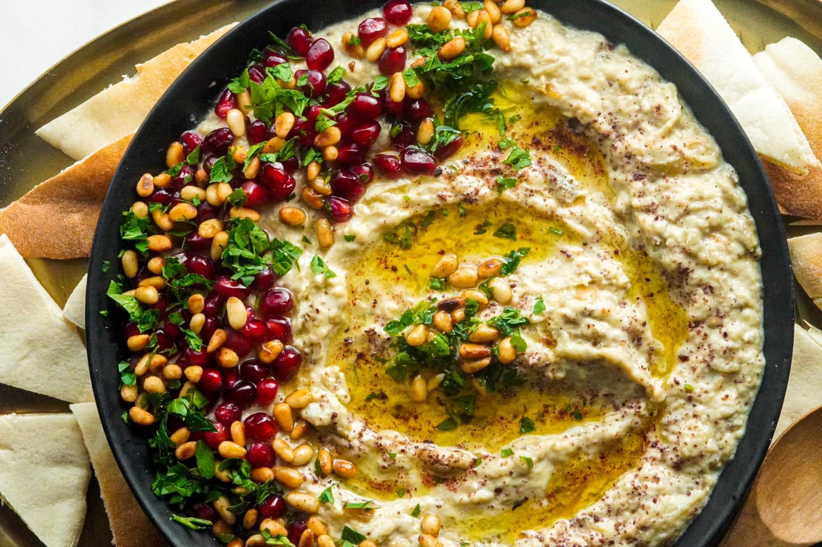 A large plate of Baba Ghanoush, topped with olive oil in the middle, pine nuts, pomegranate seeds, and parsley. Pita bread around it.