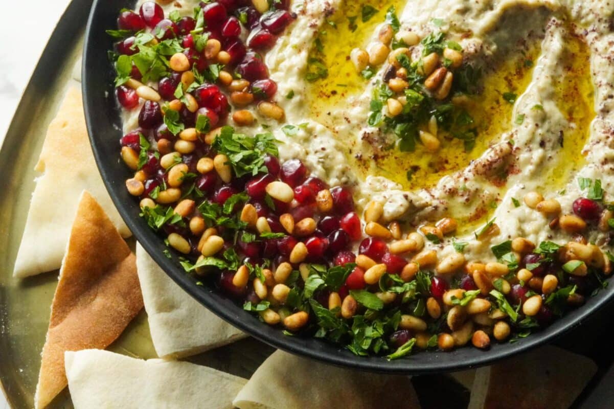 A large plate of Baba Ghanoush topped with pine nuts, pomegranate seeds, parsley, and olive oil. Serve it with triangles of Pita bread around it.