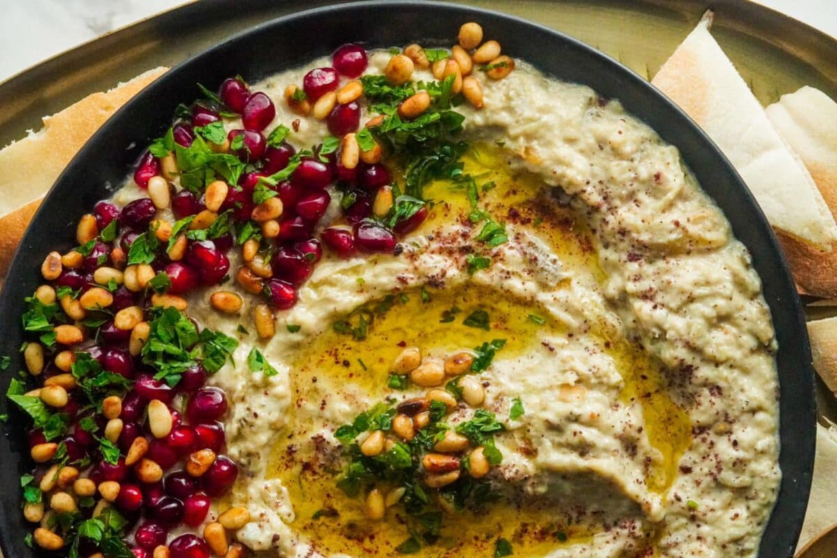 A tray with pita bread triangles and a plate of Baba Ghanoush drizzled with olive oil and topped with pine nuts, pomegranate seeds, and parsley.