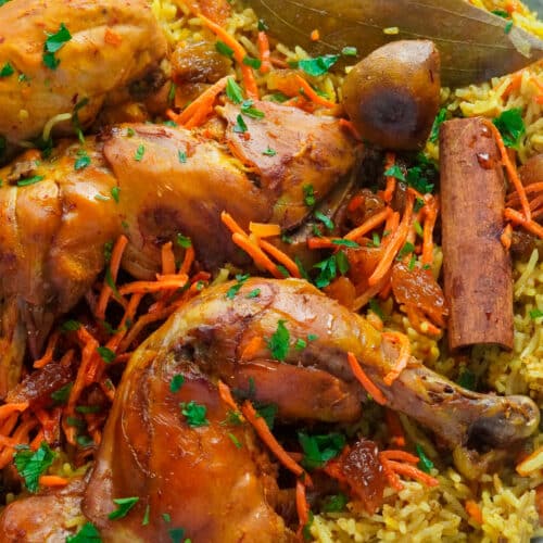 Yellow Saudi rice with well-seasoned thighs on top garnished with dried bay leaves, dry lemon, shredded carrots, and raisins.