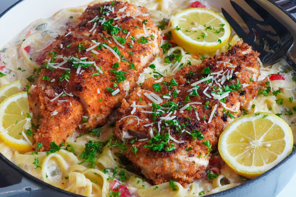 a bowl of creamy Cajun alfredo pasta served with crispy and spicy chicken cutlet on top, decorated with lemon wedges, shredded parmesan cheese, and fresh parsley