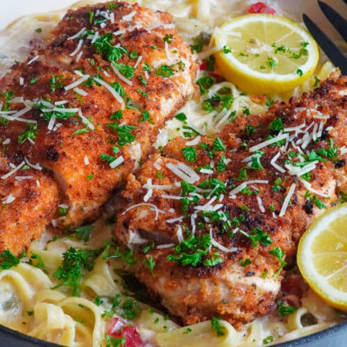 a bowl of creamy Cajun alfredo pasta served with crispy and spicy chicken cutlet on top, decorated with lemon wedges, shredded parmesan cheese, and fresh parsley