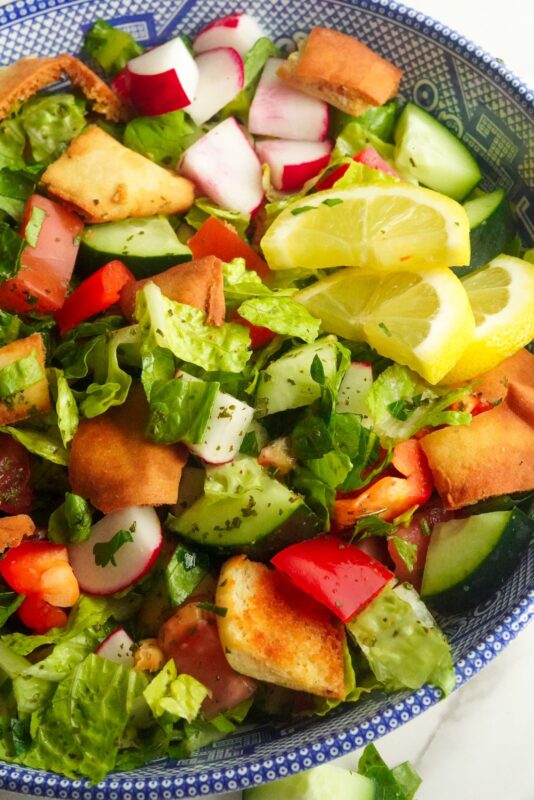 The best fattoush salad containing chopped veggies, lemon wedges and pita chips drizzled with pomegranate molasses