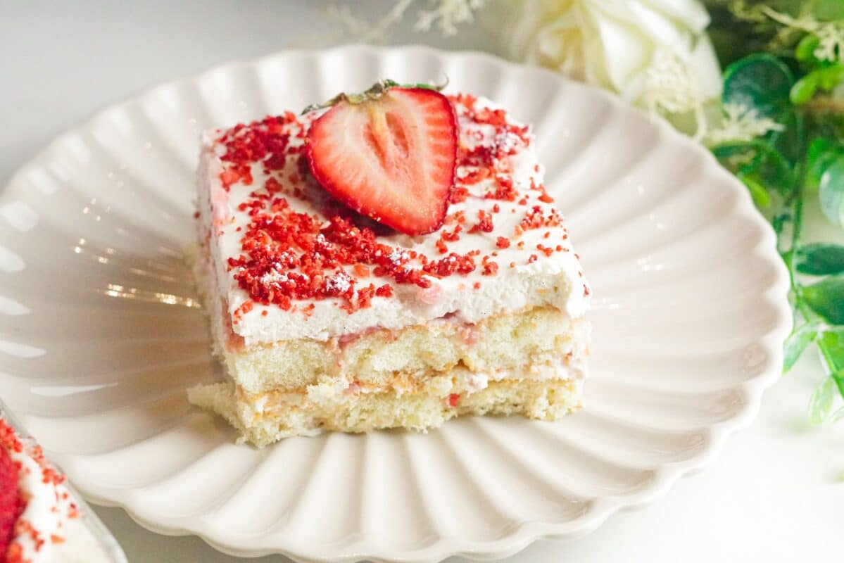 a plate contains a piece of fresh strawberry no bake tiramisu topped with a layer of vanilla-flavored mascarpone cream, crushed freeze-dried strawberries, and a half of fresh strawberry