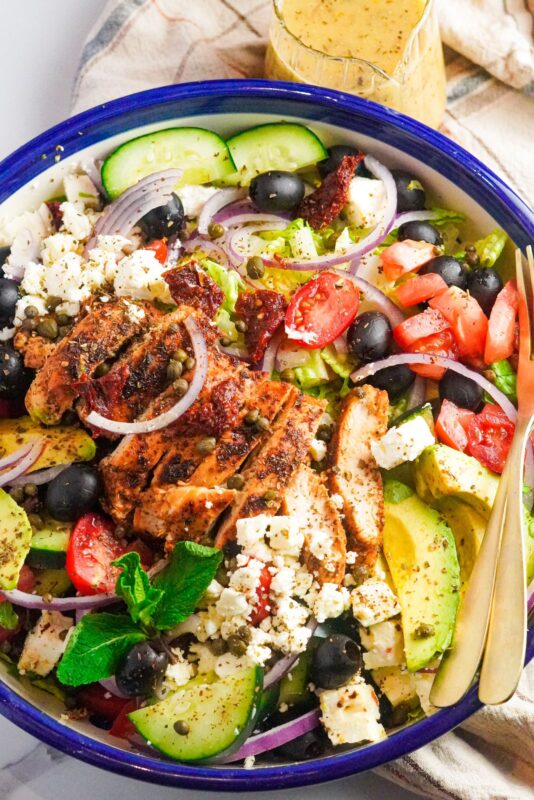 A bowl of healthy Greek Salad with Chicken made that consists of chicken breasts sliced into strips, cherry tomatoes, black olives, feta cheese, slices of onions and cucumbers and topped with  flavorful spices. 