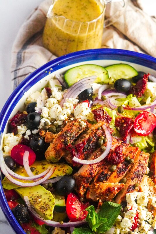 A bowl of spicy chicken, cherry tomatoes, black olives, feta cheese, avocado and cucumbers alongside a jar of tasty sauce. 