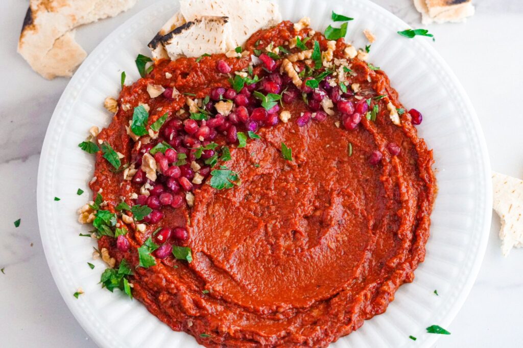 Muhammara dip in a white plate topped with chopped walnuts, pomegranate seeds, and chopped parsley and served with pita bread