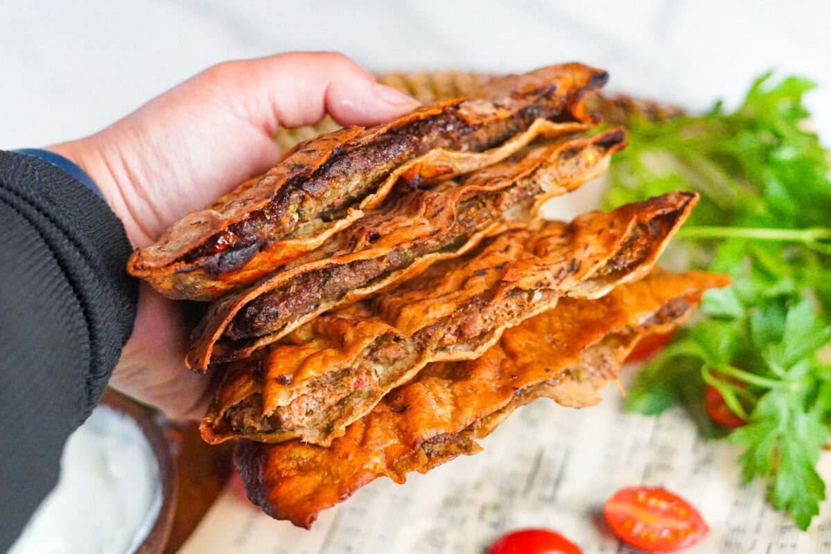 A hand holding 4 juicy pockets of ground beef stuffed pitas. They look crispy and delicious with cherry tomatoes and vibrant green parsley in the background. 