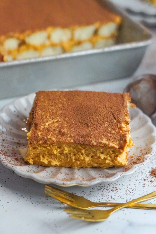 A square piece of pumpkin tiramisu is set on a plate with a sea-shell shape. It is garnished with a thin coat of ground cinnamon and cocoa.