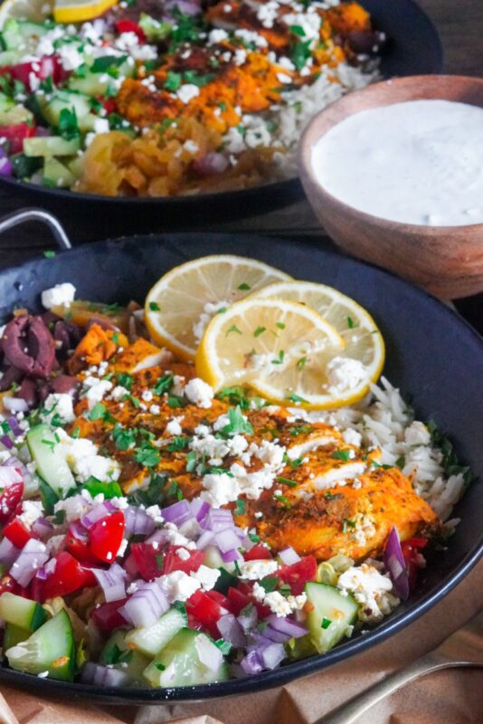 A bowl that is layered with rice, veggies, marinated and cooked chicken, feta cheese, and tzatziki sauce.