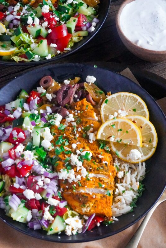 This healthy Greek chicken and rice bowl meal is served with feta cheese and lemon wedges on top and tzatziki sauce aside.