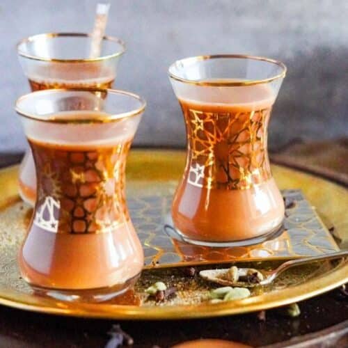 A wooden spoon leans beside a bronze tray. On top of the tray, there are 3 Arabic glasses containing Yemeni tea.