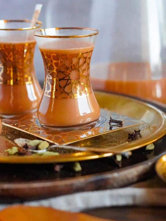 Two glasses are put on a tray. One glass has a teaspoon in it while both cups have Adeni chai.