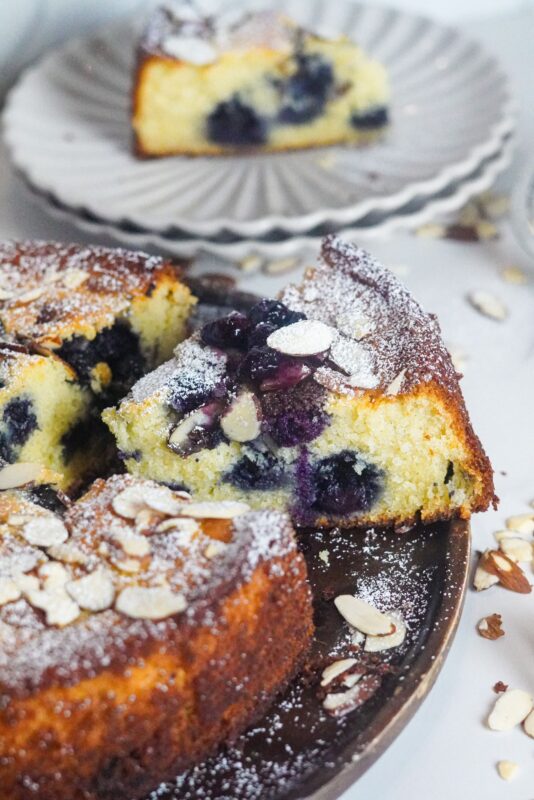 a slice cut of ricotta cake stuffed with frozen blueberries and beautifully garnished with almonds and a sprinkle of powdered sugar