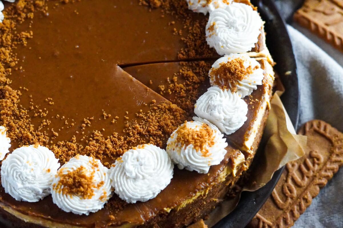 This image shows Biscoff and pumpkin cheesecake, perfectly glazed with Biscoff spread, dusted with biscoff cookie crumbles, and decorated with perfect balls of whipped cream. One perfectly triangular slice awaits being consumed.