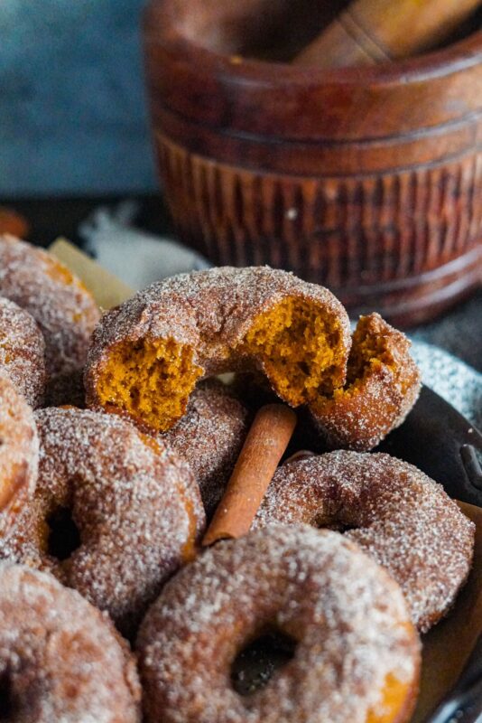 A bit fall-flavored donut next to a batch of baked donuts coated in cinnamon sugar with cinnamon sticks on the side