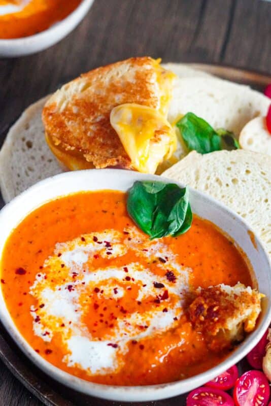 a medium bowl of roasted tomato bisque served with slices of grilled cheese bread alongside roasted garlic bulb, Roma tomatoes, and a sprinkle of Aleppo pepper garnished with fresh basil leaves