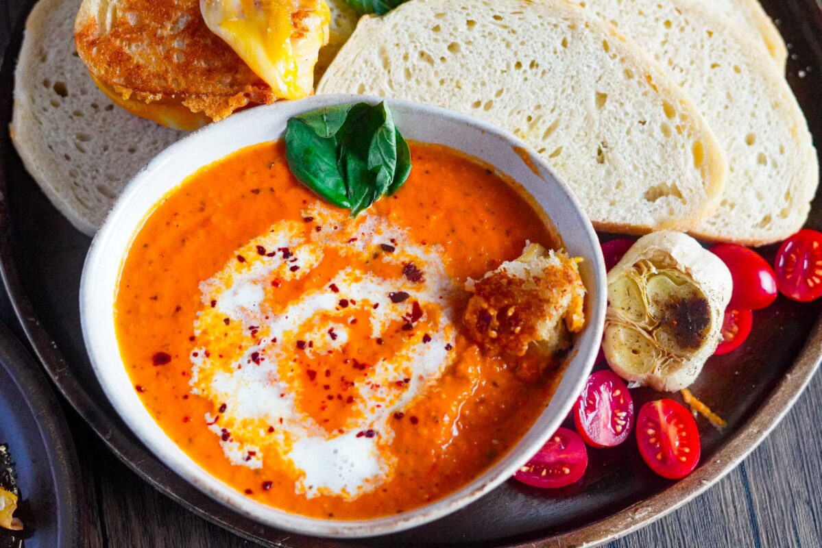 a bowl of cozy roasted tomato bisque served with slices of French bread alongside roasted garlic bulb, Roma tomatoes, and a sprinkle of Aleppo pepper garnished with fresh basil leaves