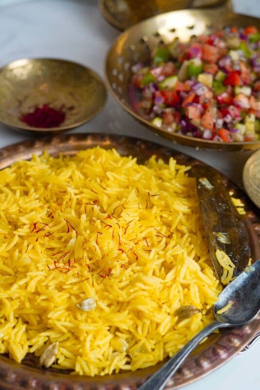 Yellow saffron rice, garnished with vibrant red saffron threads and a bay leaf, is set within fancy, traditional copper plates with a salad on the side.