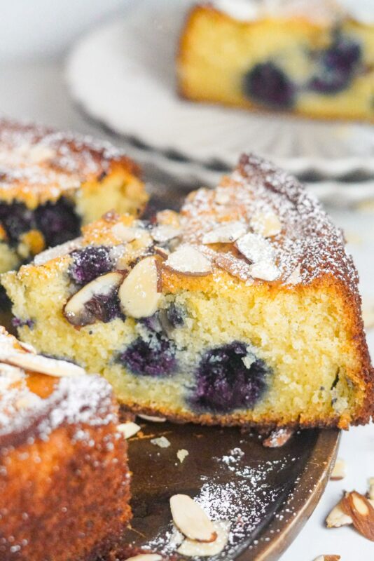A close-up shot capturing a slice of Blueberry Ricotta cake adorned with almonds on its surface and a sprinkle of powdered sugar.