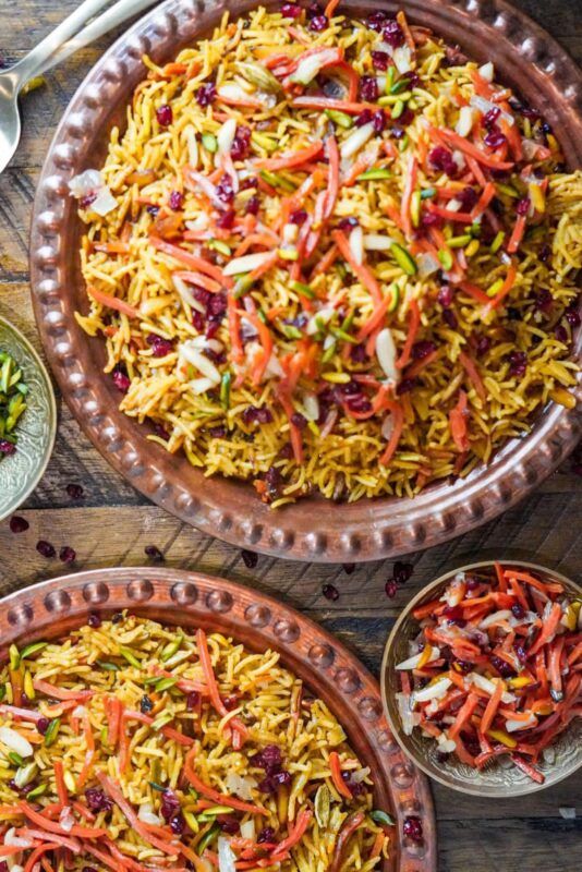 A rich bowl of Iranian rice topped with pistachios and carrots shreds on a wooden table surrounded by another plate and a side plate of the toppings.