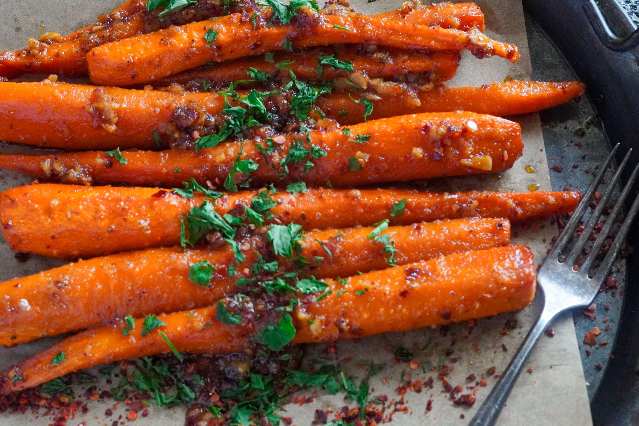 A lined pan of carrots seasoned with honey, garlic, olive oil, and other seasonings and topped with some chopped parsley