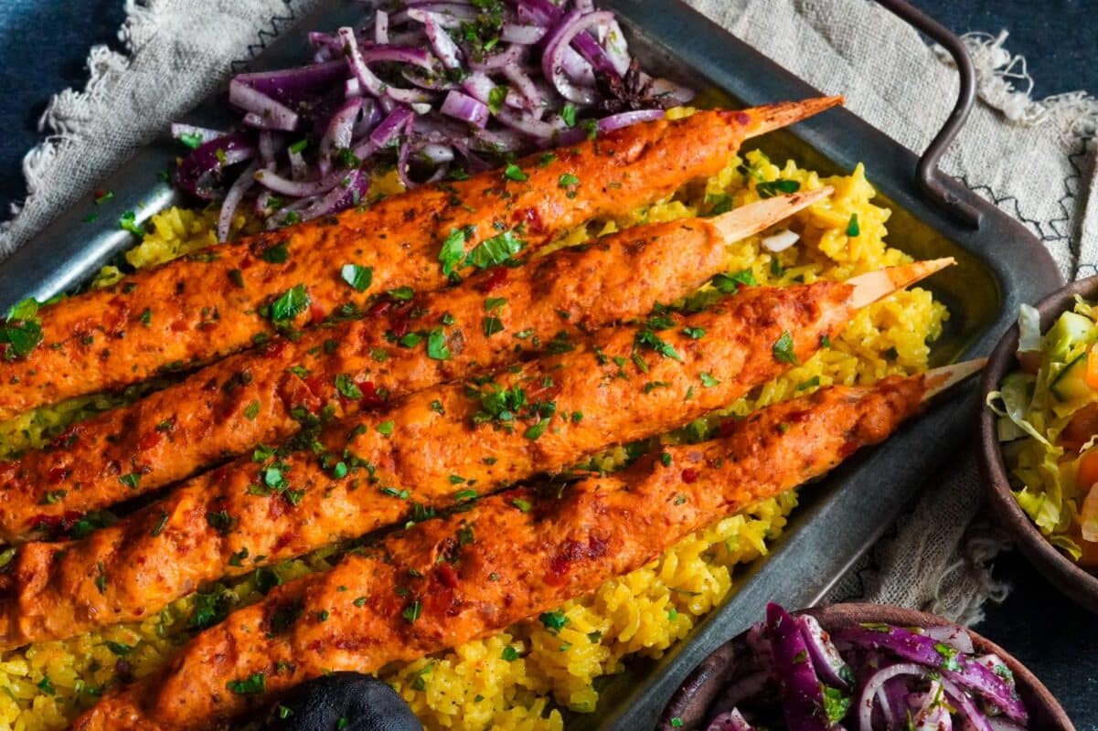 four chicken kafta skewers sprinkled with finely chopped parsley and served with sumac onion salad on a bed of yellow rice