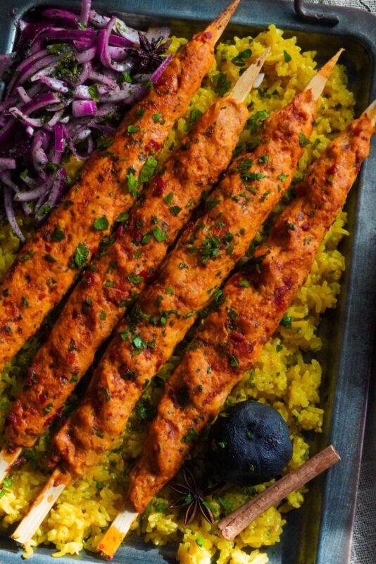 four baked chicken kafta skewers sprinkled with finely chopped parsley and served with sumac onion salad on a bed of yellow rice and dried lime