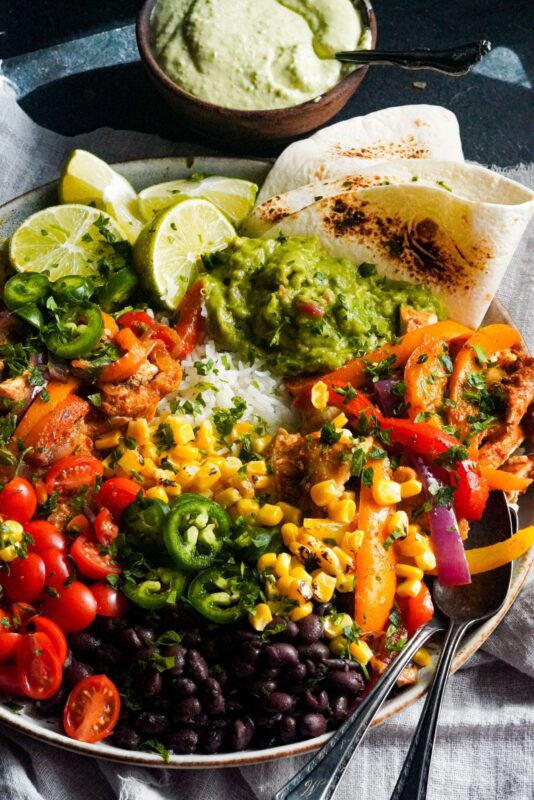 delicious bowl of chicken fajitas on a base of rice and topped with tomatoes, guacamole, corn, beans, lime wedges, and a sprinkle of chopped cilantro
