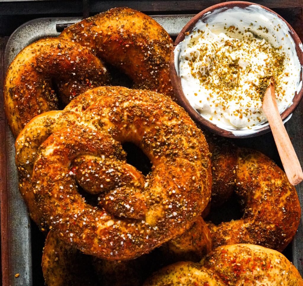 A golden-brown zaatar pretzels sprinkled with flakey salt alongside a bowl of labneh.