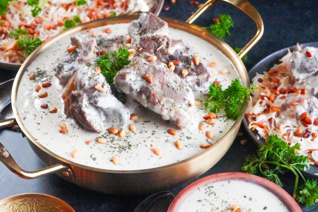 A large golden bowl of laban immo stew made up of lamb shanks with warm yogurt soup. The bowl is topped with fried nuts and sprigs of fresh cilantro.