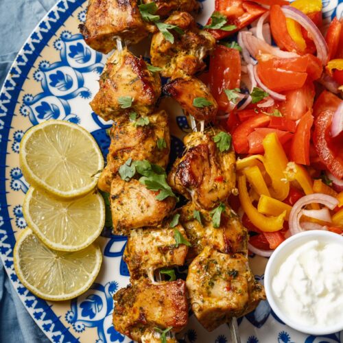 a close view of a plate of two skewers of shish tawook grilled chicken sprinkled with finely chopped parsley served with Arabic chopped salad, a small bowl of garlic sauce, and lemon wedges