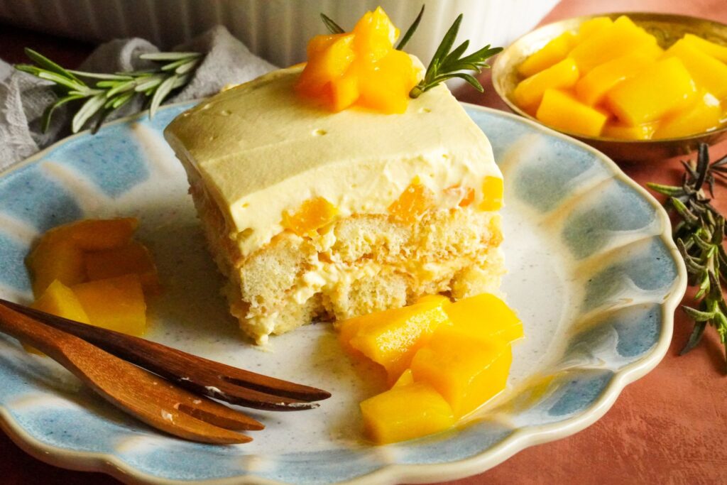 A close-up view of a mango tiramisu slice is served on a plate and topped with mango chunks and rosemary spring.