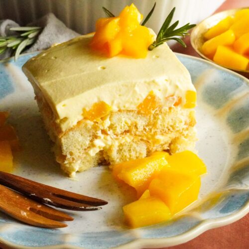 A close-up view of a mango tiramisu slice is served on a plate and topped with mango chunks and rosemary spring.