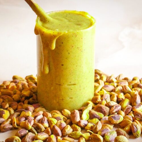 A tall glass filled to the brim with green pistachio spread sits amidst a bunch of pistachios, with the spread dripping down its sides.