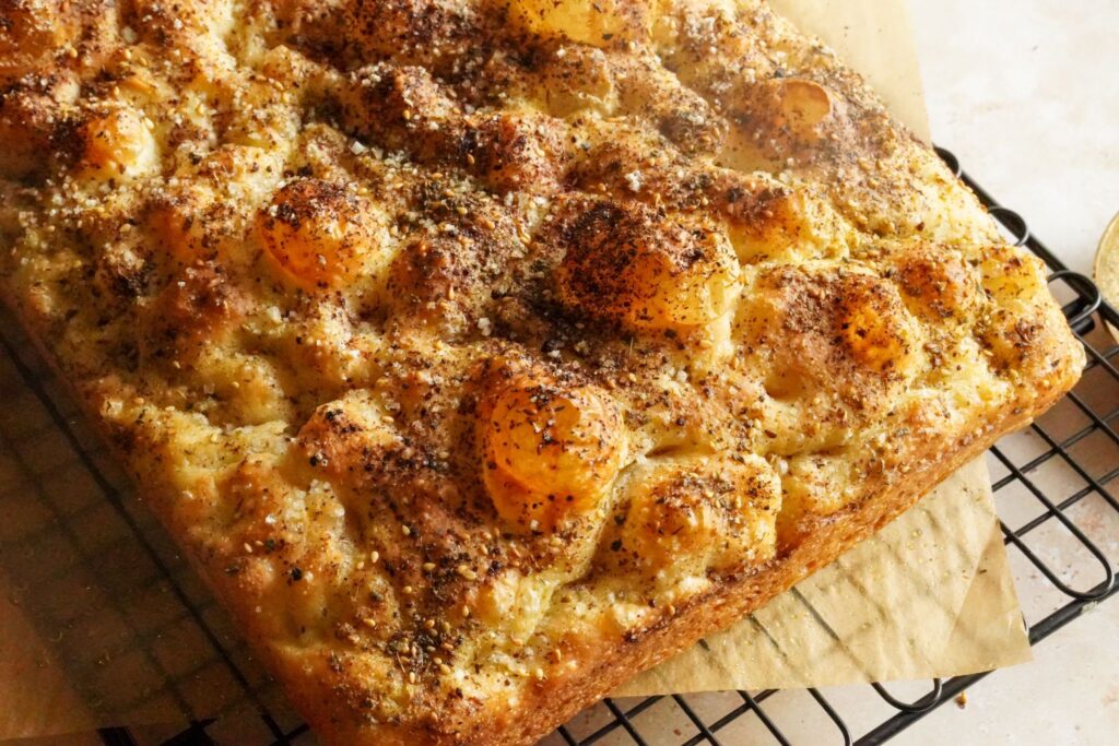 A golden crispy loaf of focaccia bread topped with zaatar and sumac, placed on a baking rack.