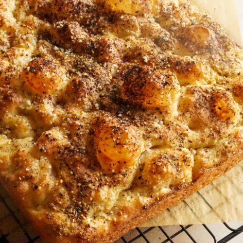 A golden crispy loaf of focaccia bread topped with zaatar and sumac, placed on a baking rack.